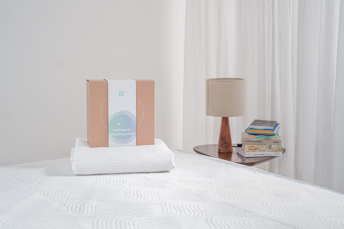 hộp giấy nằm trên chiếu điều hoà cho bé gắp gọn trên nệm, bên phải là bàn cạnh giường có đèn ngủ và chồng sách