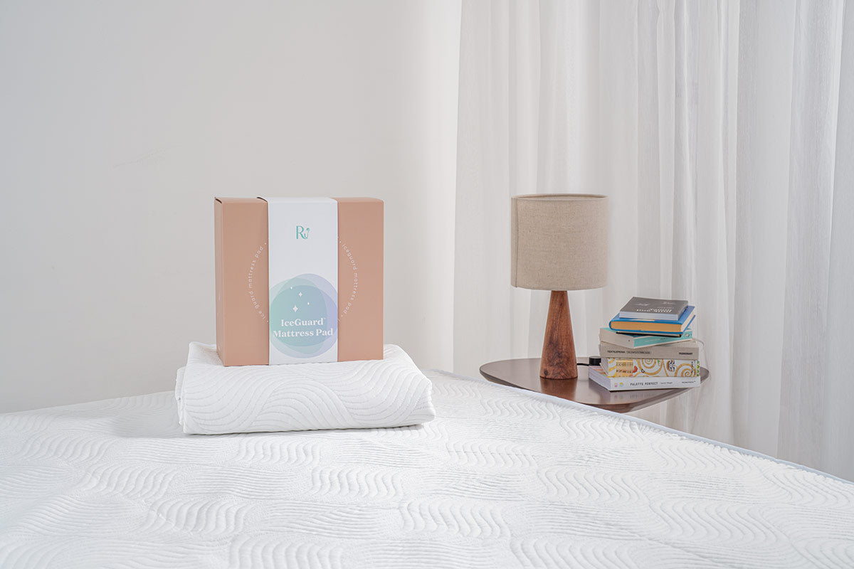 hộp carton đặt trên chiếu điều hoà có công nghệ làm mát tự nhiên gấp gọn đặt lên giường, bên phải là đèn ngủ và chồng sách trên bàn để đầu giường