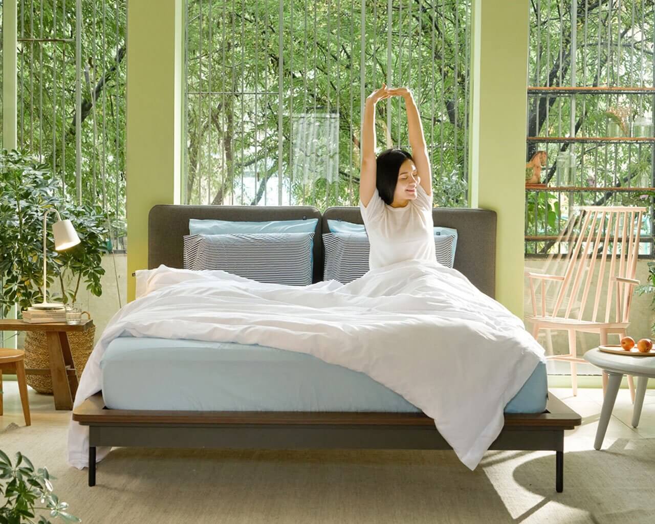 cô gái thức dậy vào buổi sáng, nửa người vẫn còn đang ở trong chăn, bao quanh căn phòng là nhiều cây xanh