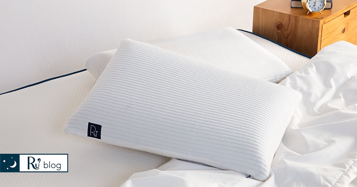 Chiếc gối nằm tựa lên một chiếc gối khác và mền trên giường, cạnh bên là nightstand màu gỗ
